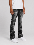 A person wearing distressed black and gray denim jeans with silver studs and ripped details, paired with a white t-shirt and white sneakers, shown from the waist down on a light gray studio background.
