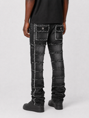 Rear view of a person wearing black patchwork denim jeans with frayed seams and stacked patch pockets, paired with a black t-shirt and black shoes, on a light gray studio background.
