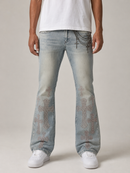A person wearing light-wash blue jeans with large rhinestone-embellished cross motifs on the lower legs, paired with a white t-shirt and white sneakers, shown from the waist down against a plain light beige studio background.